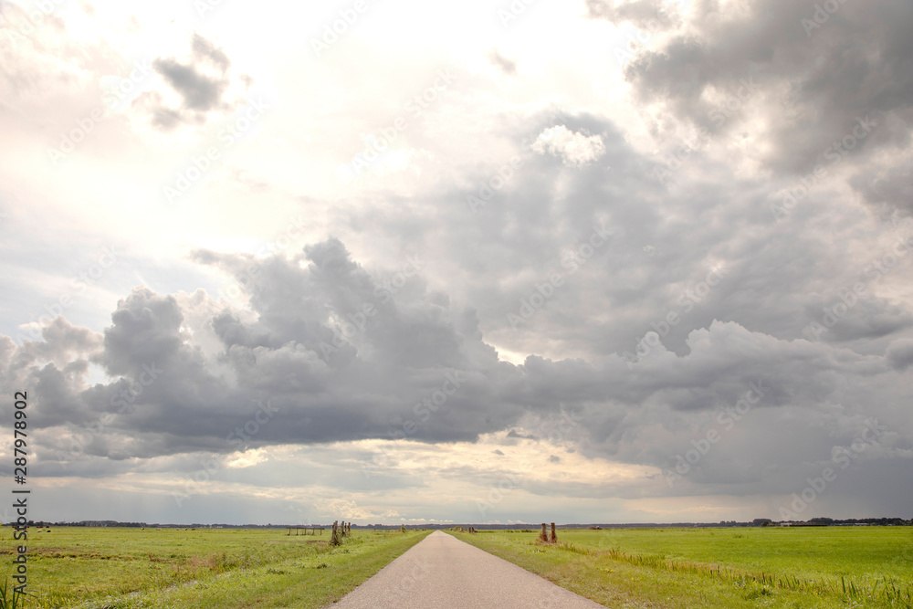 Road in Holland countryside, perspective, under heavy dark cloudy skies and between meadows and a faraway straight horizon. Road in Holland countryside, perspective, under heavy dark cloudy skies and between meadows and a faraway straight horizon.