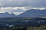 Panoramic view of an extensive spring green valley with a lake and a small town with mountains and clouds in the background