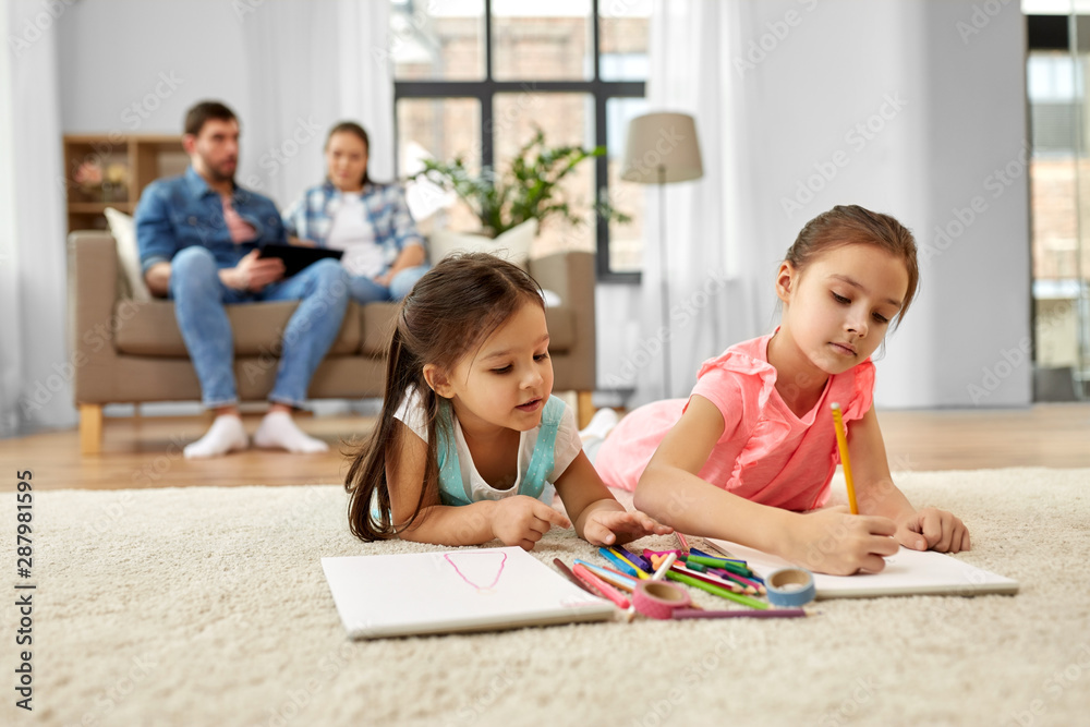 family, leisure and childhood concept - happy sisters lying on floor and drawing in sketchbooks with crayons at home family, leisure and childhood concept - happy sisters lying on floor and drawing in sketchbooks with crayons at home