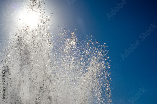 jets and drops of water of the fountain on the background of the sun and blue sky with sun flare