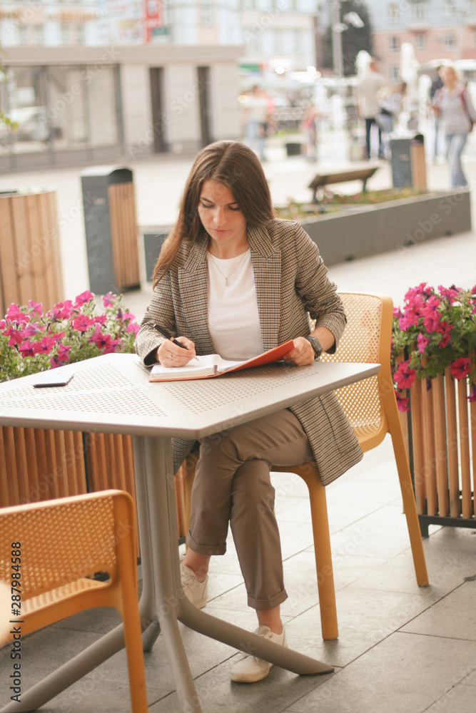 A woman with long dark hair in a jacket and sneakers sits at a table in a street cafe and writes in a diary