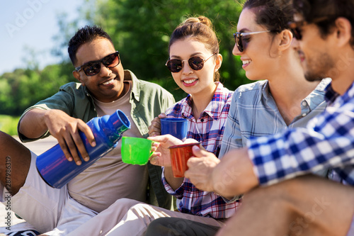 leisure, picnic and people concept - happy friends drinking tea from thermos outdoors in summer park