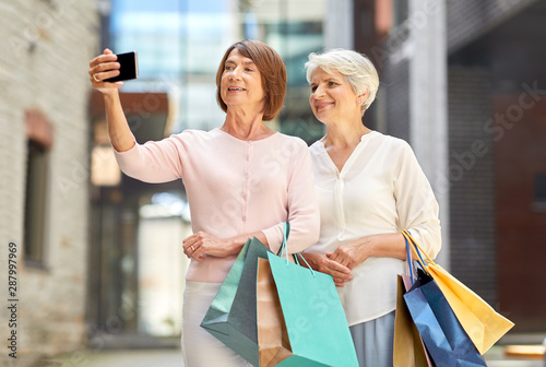 sale, consumerism and people concept - two senior women or friends with shopping bags taking selfie by smartphone on tallinn city street photo