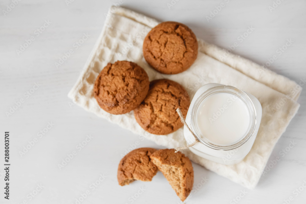 Milk in glass jar and oatmeal cookies near napkin on white table background. Top view. Copy, empty space for text Milk in glass jar and oatmeal cookies near napkin on white table background. Top view. Copy, empty space for text