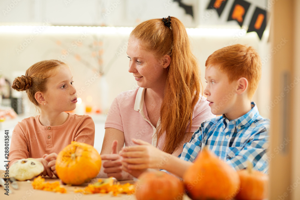 Smiling young mother sitting at the table and talking to her two children while they making crafts from pumpkins together at home Smiling young mother sitting at the table and talking to her two children while they making crafts from pumpkins together at home