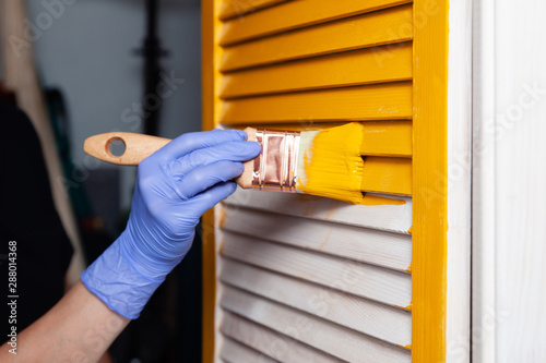 Closeup woman hand in purple rubber glove with paint brush painting natural wooden door with yellow paint, creative design house renovation theme. How to Paint Wooden Surface. Selected focus
