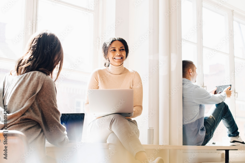 Smiling woman looking away while sitting with colleagues at desk by window in office Smiling woman looking away while sitting with colleagues at desk by window in office