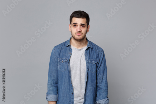 Handsome stylish unshaven young man in denim jeans shirt posing isolated on grey wall background studio portrait. People sincere emotions lifestyle concept. Mock up copy space. Looking camera sceptic.