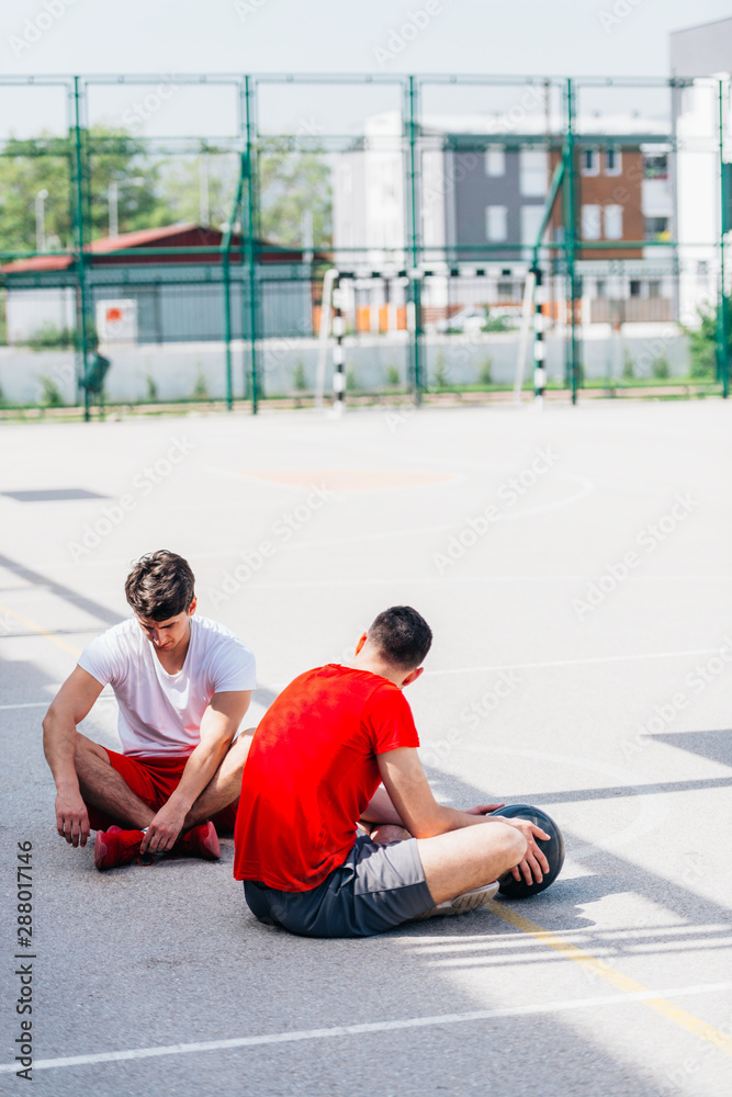 Two strong caucasian athletes resting on the ground at the basketball court while having a conversation. Two strong caucasian athletes resting on the ground at the basketball court while having a conversation.