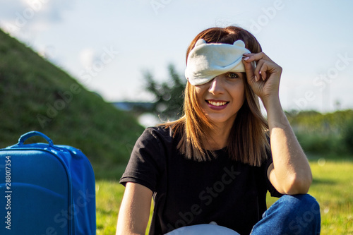 Young smiling girl with suitcase and sleeping mask on her face. Jet lag concept.