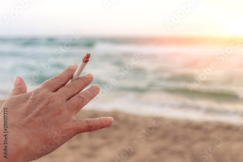 Hand holds a cigarette against the evening sea. © Oleg