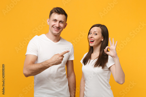 Young couple friends guy girl in white empty blank design t-shirts posing isolated on yellow orange background. People lifestyle concept. Mock up copy space. Showing OK gesture, pointing finger aside.