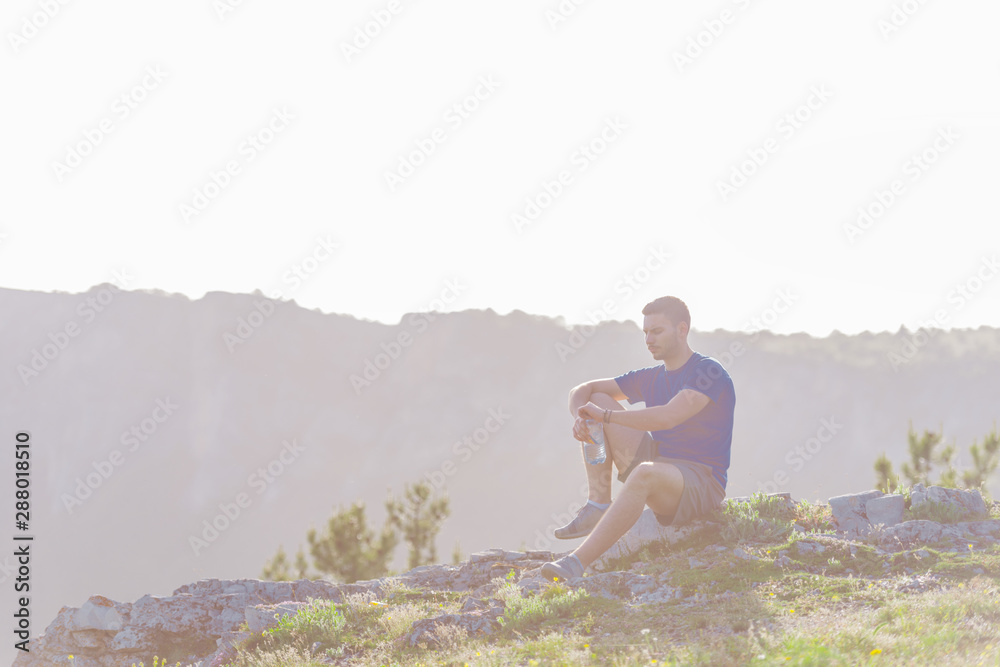 Handsome athlete sitting at a rocky peak while looking at the breathtaking mountain line and a beautiful lake while wearing a blue shirt and grey shorts.