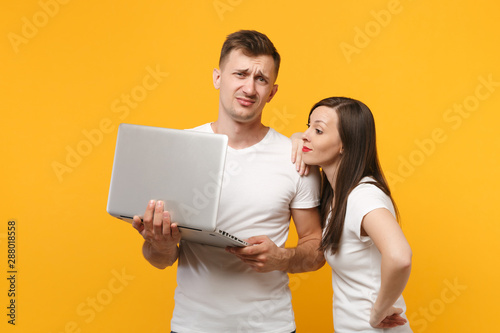 Displeased young couple two friends guy girl in white blank t-shirts posing isolated on yellow orange background. People lifestyle concept. Mock up copy space. Holding, working on laptop pc computer.