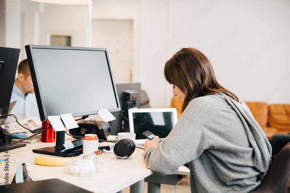 Side view of female programmer using mobile phone on desk while sitting in office Side view of female programmer using mobile phone on desk while sitting in office