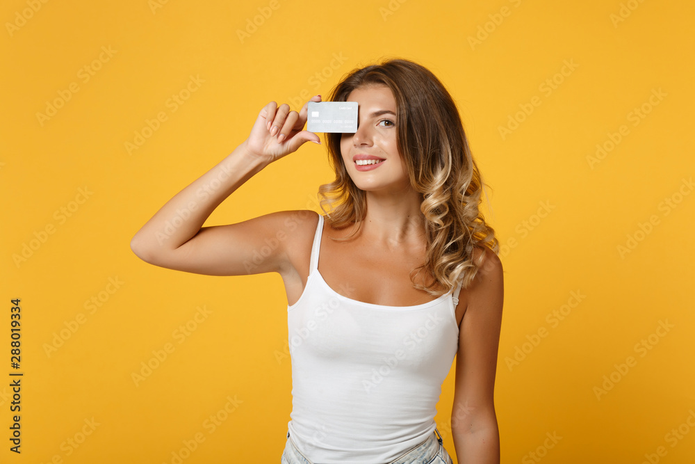 Smiling young woman girl in light casual clothes posing isolated on yellow orange wall background, studio portrait. People lifestyle concept. Mock up copy space. Covering eye with credit bank card. Smiling young woman girl in light casual clothes posing isolated on yellow orange wall background, studio portrait. People lifestyle concept. Mock up copy space. Covering eye with credit bank card.