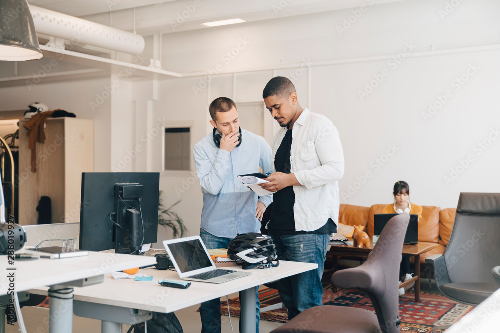 Male IT professionals discussing over digital tablet while standing against wall in creative office Male IT professionals discussing over digital tablet while standing against wall in creative office