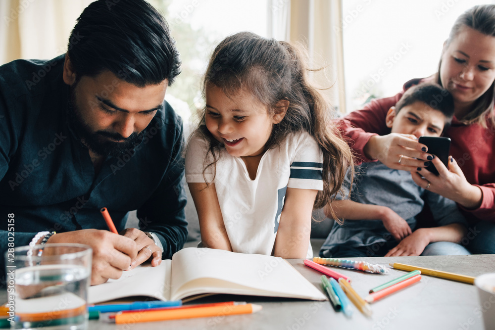 Father teaching drawing to daughter while woman using smart phone with autistic son in background Father teaching drawing to daughter while woman using smart phone with autistic son in background