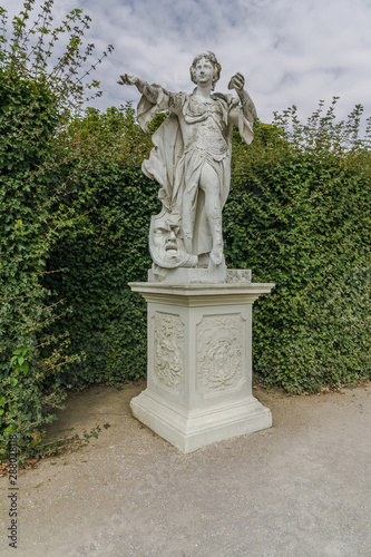 Female Statue in beautiful garden. Monument made of white stone stands on rectangular pedestal. Statue of woman which holds out one of her hand and the othe hand holds a shell