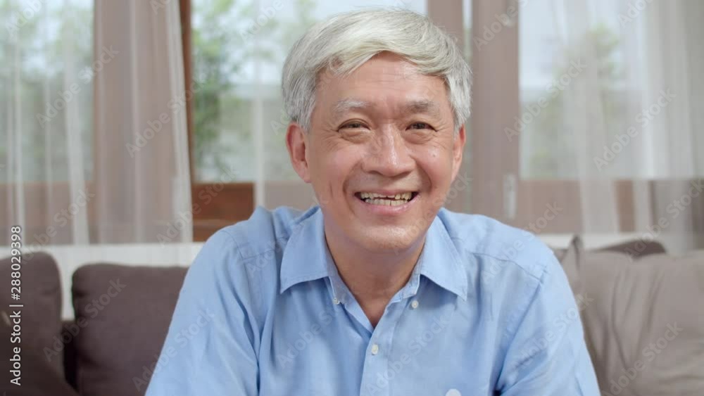 Asian senior man video call at home. Asian senior older Chinese male using mobile phone video call talking with family grandchild kids while lying on sofa in living room at home. Looking at camera. Asian senior man video call at home. Asian senior older Chinese male using mobile phone video call talking with family grandchild kids while lying on sofa in living room at home. Looking at camera.