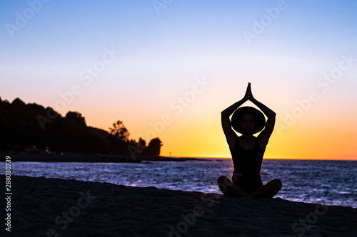 Woman meditating and doing yoga on the beach during sunset with her hat in lotus position
