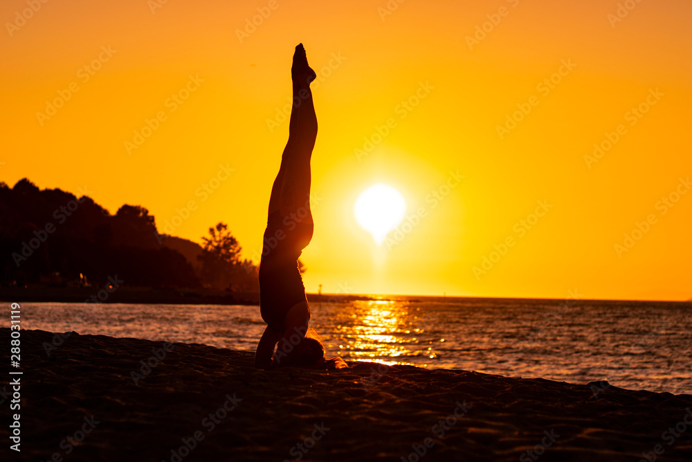 Flexible woman practicing yoga and gymnastics on the beach at sunset in balanced arm pose, wellbeing, good posture, zen attitude, healthy, pacific ocean view and orange sky