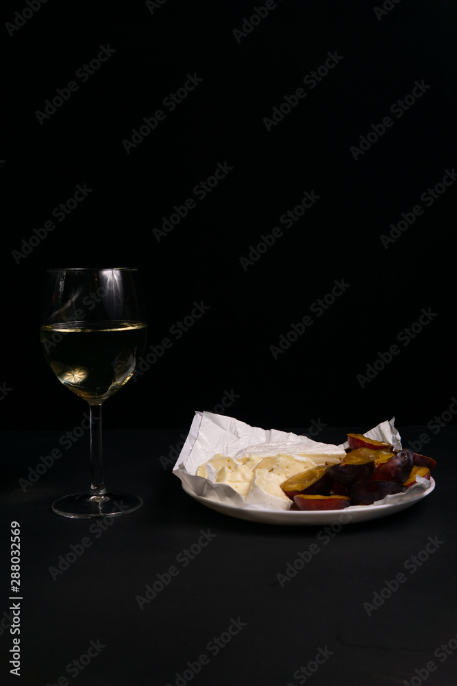 glass of wine next to a slice of cheese, plums on a black background