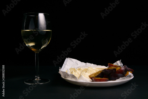 glass of wine next to a slice of cheese, plums on a black background