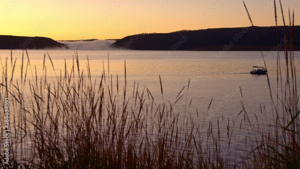 Slow motion of boat trolling on lake during sunrise moving on slider looking through tall grass. Slow motion of boat trolling on lake during sunrise moving on slider looking through tall grass.
