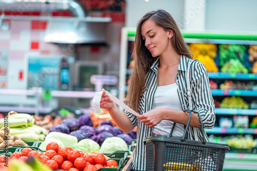 Obraz na plátně Young woman with shopping basket checks and examines a sales receipt after purchasing food in a grocery store