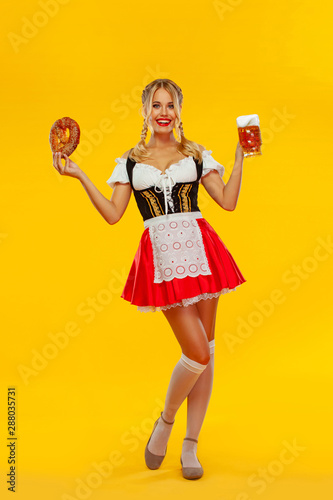 Young sexy Oktoberfest girl waitress, wearing a traditional Bavarian or german dirndl, serving big beer mugs with drink and bretzel, isolated on yellow background. Full height photo