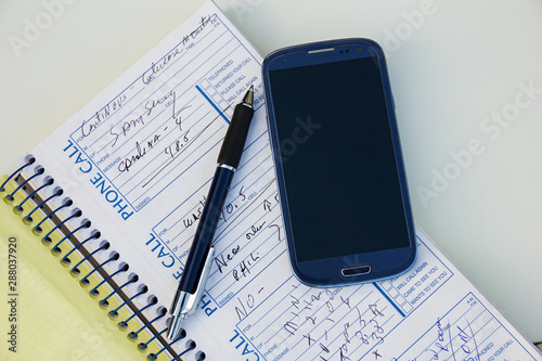 Smartphone, pen, hand, note book or phone book or organizer with some notes, info or phones number on a white table. Plans and agenda. Keyboard on.