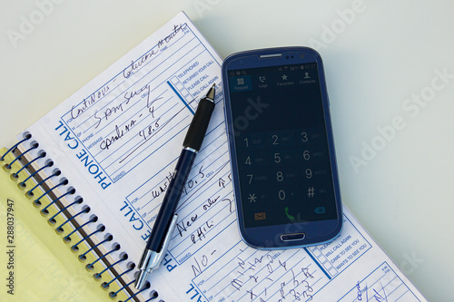 Smartphone, pen, hand, note book or phone book or organizer with some notes, info or phones number on a white table. Plans and agenda. Keyboard on.
