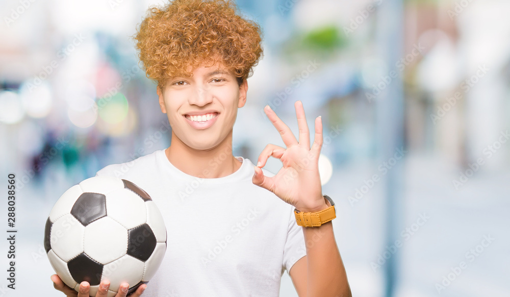 Young handsome man holding soccer football ball doing ok sign with fingers, excellent symbol Young handsome man holding soccer football ball doing ok sign with fingers, excellent symbol