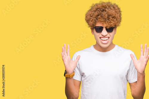 Young handsome man with afro hair wearing sunglasses crazy and mad shouting and yelling with aggressive expression and arms raised. Frustration concept.
