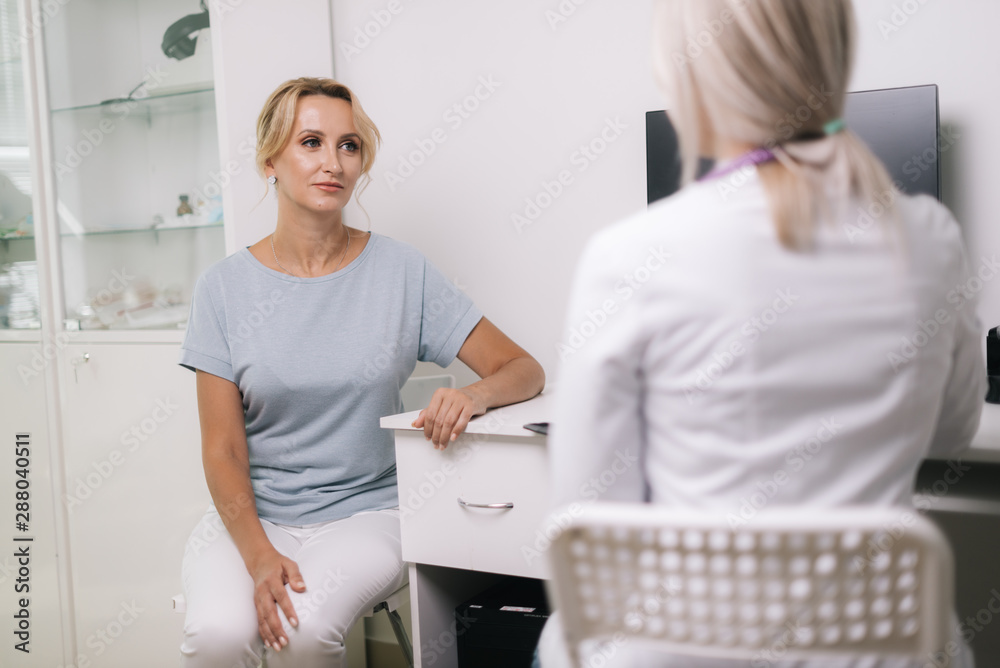 Fototapeta premium Young blonde woman at doctor checkup in white dressing gown. Therapist female sitting at table and asks the patient questions