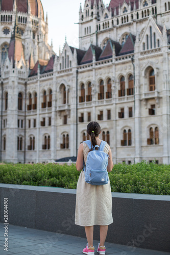 budapest travel: brunette tourist girl with a backpack in a dress stands near the Hungarian Parliament 1