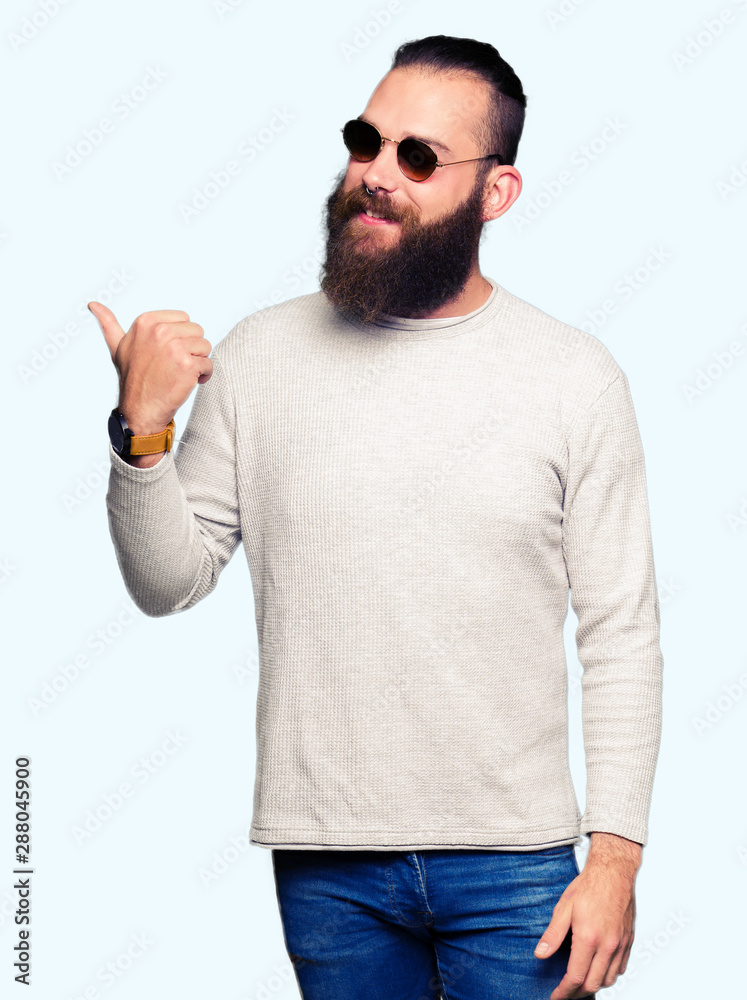 Young hipster man wearing sunglasses smiling with happy face looking and pointing to the side with thumb up. Young hipster man wearing sunglasses smiling with happy face looking and pointing to the side with thumb up.