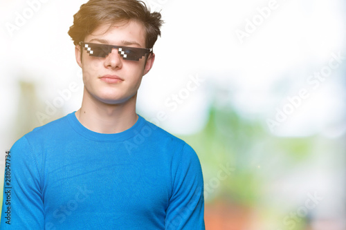 Young man wearing funny thug life glasses over isolated background with serious expression on face. Simple and natural looking at the camera.