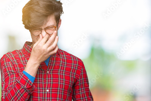Young handsome man wearing glasses over isolated background smelling something stinky and disgusting, intolerable smell, holding breath with fingers on nose. Bad smells concept.