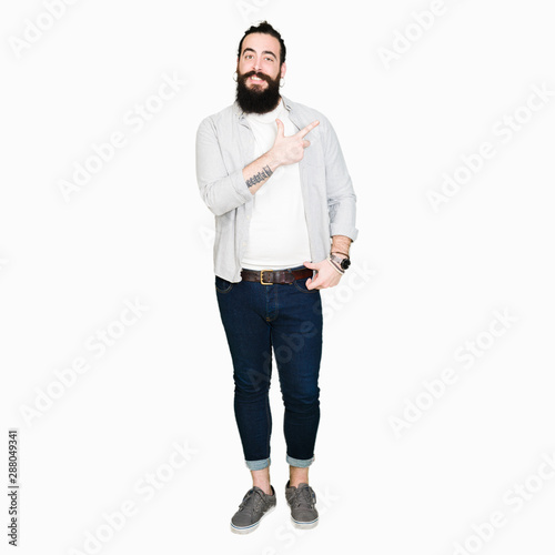 Young man with long hair, beard and earrings cheerful with a smile of face pointing with hand and finger up to the side with happy and natural expression on face