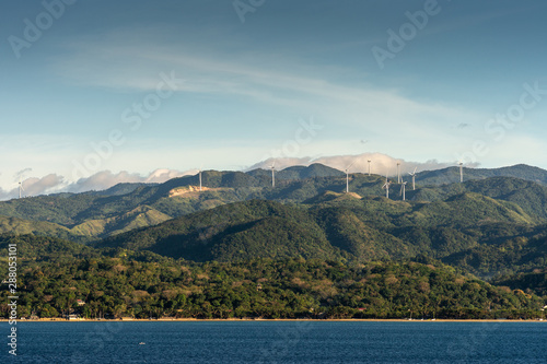 Caticlan, Malay, Philippines - March 4, 2019: Green mountain range with windmills dispersed under blue sky with thick gray cloudscape. Dark blue sea. photo
