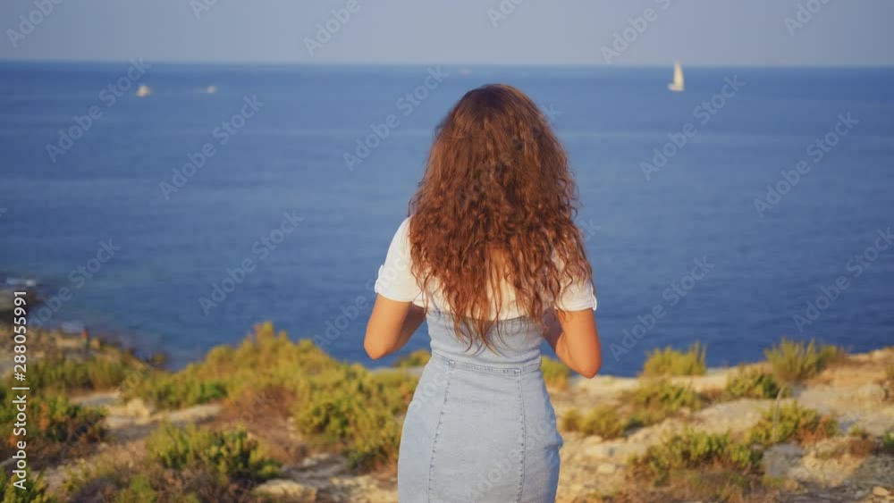 A young slender brunette woman stands on a cliff and photographs the sea and nature. Back view A young slender brunette woman stands on a cliff and photographs the sea and nature. Back view