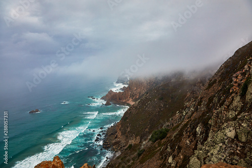 Fog envelops the rocky shore. The waves of the Atlantic Ocean break on the majestic cliffs of the westernmost cape of the Eurasian continent. Image of bad weather, poor visibility at sea.
