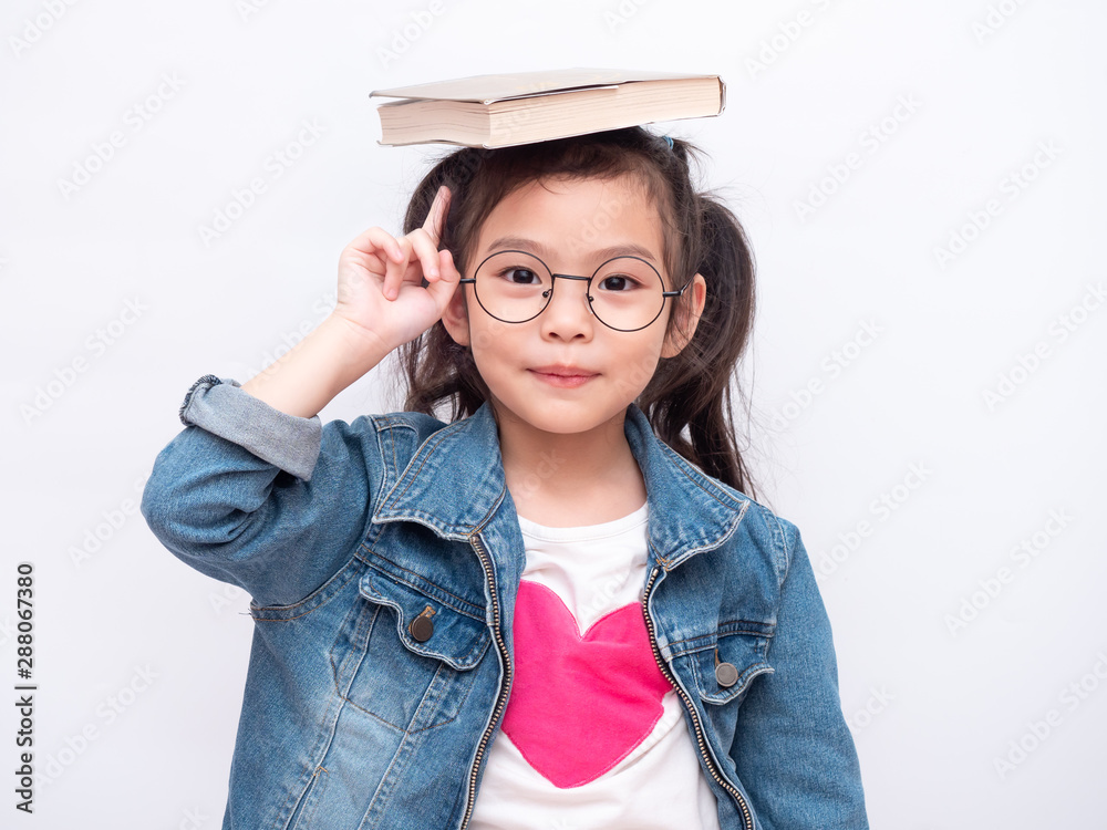 Asian little cute girl wearing glasses and put the book on head. Preschool lovely kid with the book on her head and pointing finger up. Learning and education of kid. Asian little cute girl wearing glasses and put the book on head. Preschool lovely kid with the book on her head and pointing finger up. Learning and education of kid.