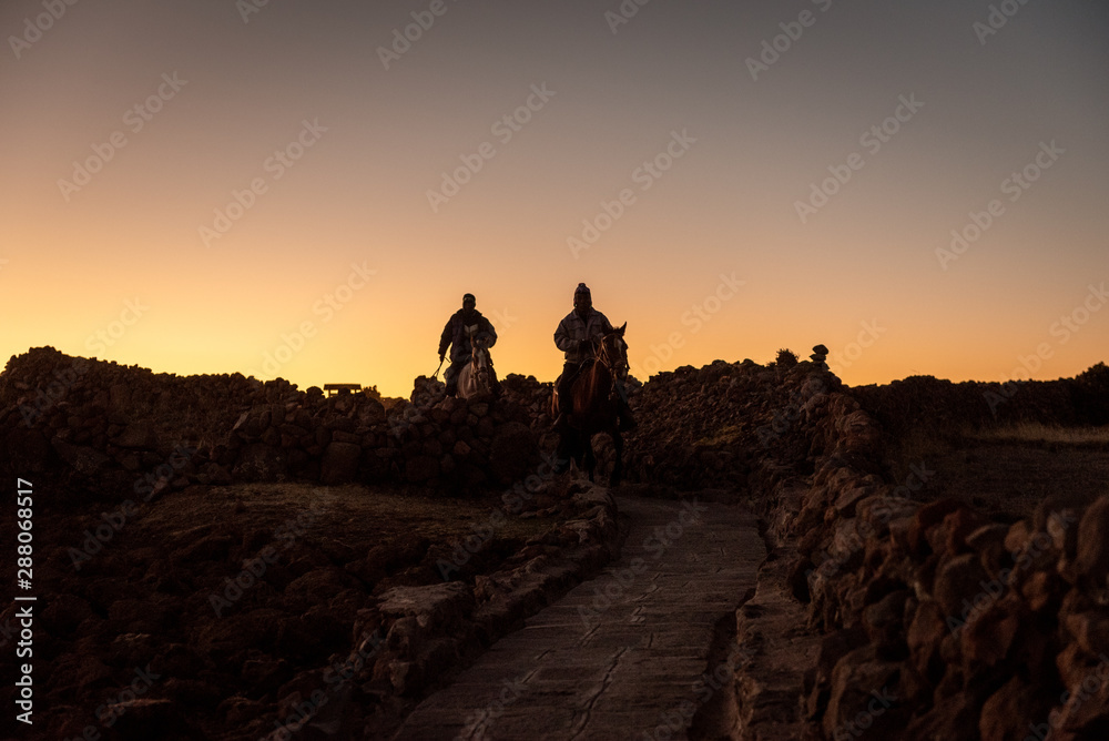 Two Peruvian horse riders following along a stone path during a cool mornings sunrise to start their long day on the farm.