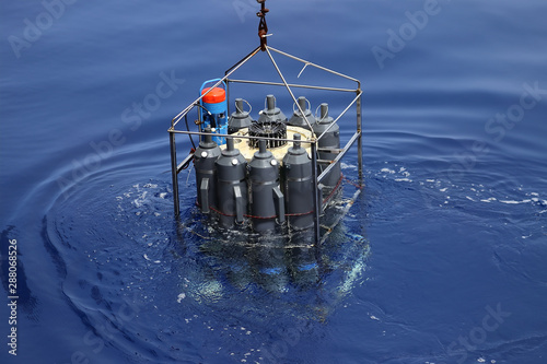 CTD rosette sampler with bathometers and other science equipment for measurement of underwater environment parameters like temperature, salinity and others. Sampler is half submerged in water.