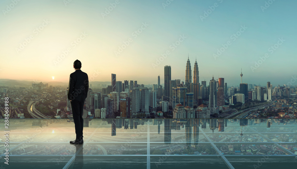 Businessman standing at transparent glass floor on rooftop with city skyline, success and thinking concept  .
