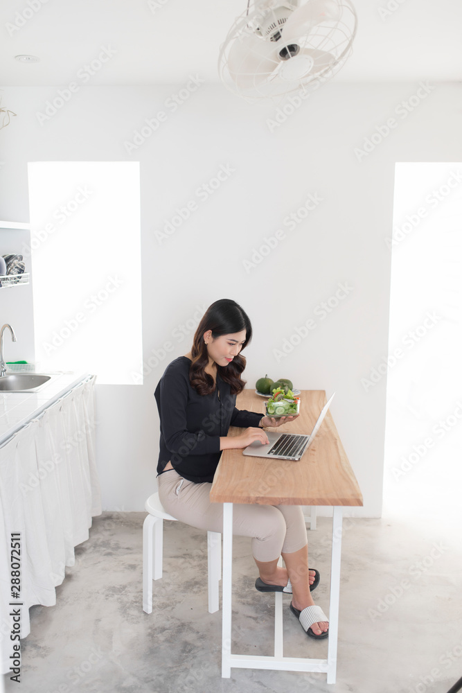 Young woman with salad working on laptop in the kitchen Young woman with salad working on laptop in the kitchen