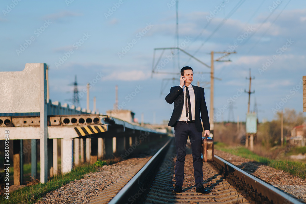 A cool and modern male businessman in a suit with a suitcase is walking along the paths from the train and talking on the phone. Business walk and serious conversation. Late for the train.